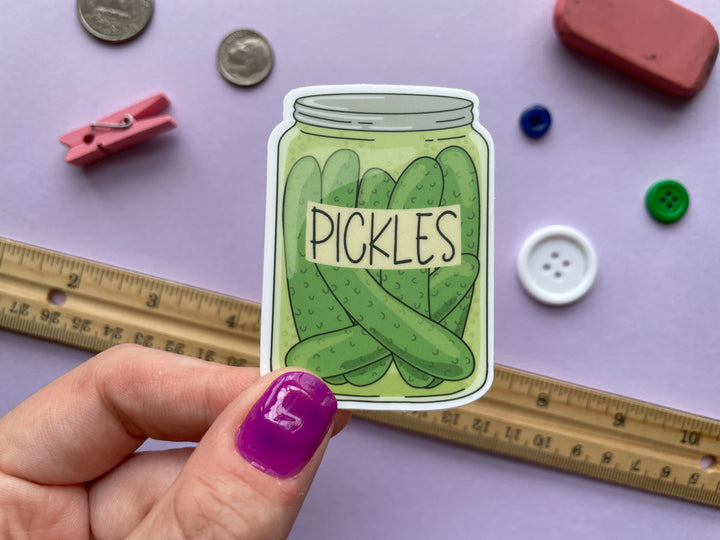 A sticker of a cartoon drawing of a jar of whole dill pickles with a label reading "pickles" is held about a purple background covered in small objects.
