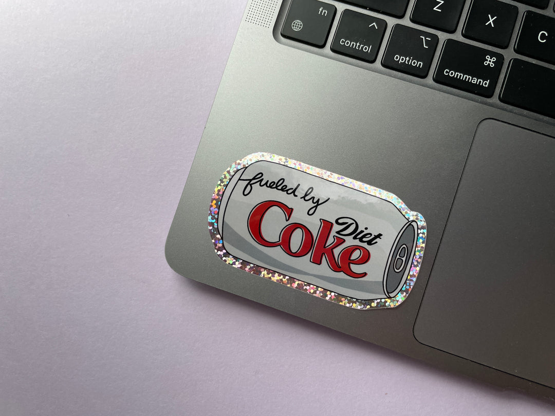A glittery sticker of a cartoon of a silver can of Diet Coke on its side with the text "fueled by" in the top corner is shown on a silver laptop.
