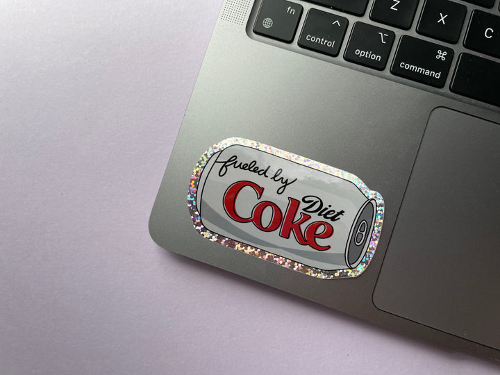 A glittery sticker of a cartoon of a silver can of Diet Coke on its side with the text "fueled by" in the top corner is shown on a silver laptop.