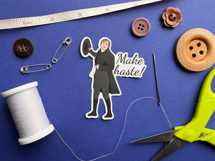 A sticker of Mr Collins from Pride and Prejudice 1995 with the text 'Make haste'. Resting on a blue background surrounded by small sewing notions.