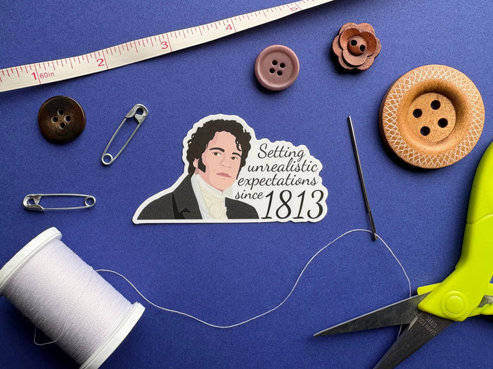A cartoon of Mr Darcy from Pride and Prejudice with the text 'Setting unrealistic expectations since 1813'. The sticker is resting on a blue background surrounded by sewing notions.