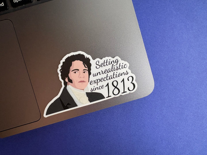 A cartoon of Mr Darcy from Pride and Prejudice with the text 'Setting unrealistic expectations since 1813'. The sticker is resting on a silver laptop on a blue background.