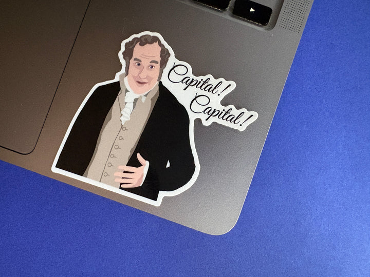 A cartoon of Sir William Lucas from Pride and Prejudice 1995 with the text 'Capital! Capital!'.  the sticker is on a silver laptop resting on a blue background.