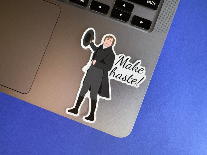 A sticker of Mr Collins from Pride and Prejudice 1995 with the text 'Make haste'. Sitting on a silver laptop resting on a blue background.