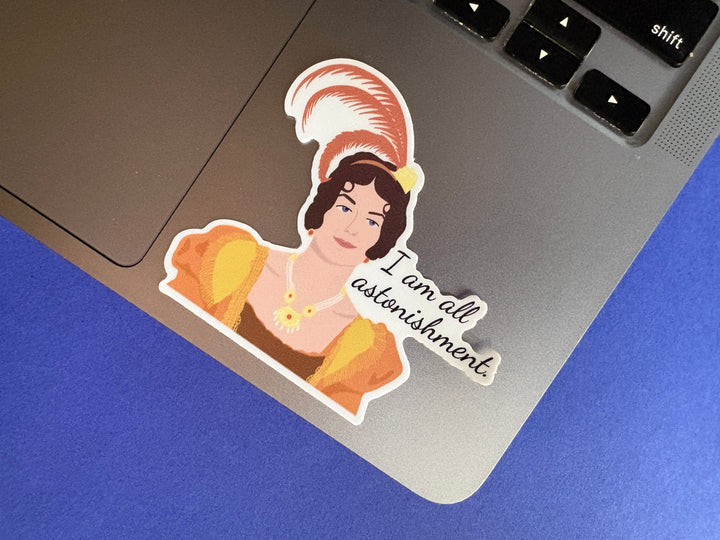 Sticker of a cartoon of Caroline Bingley from Pride and Prejudice 1995 with the text 'I am all astonishment'. Sitting on silver laptop resting on a blue background.