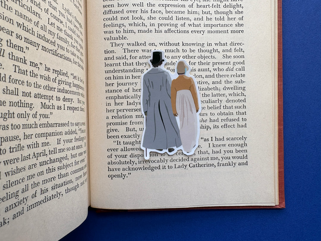 A cartoon of Mr Darcy and Elizabeth from Pride and Prejudice 1995 walking down the lane shown from behind. Shown on an open book resting on a blue background.