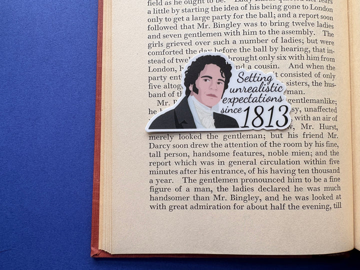 A cartoon of Mr Darcy from Pride and Prejudice with the text 'Setting unrealistic expectations since 1813'. The sticker is resting on an open book on a blue background.