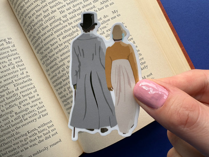 A cartoon of Mr Darcy and Elizabeth from Pride and Prejudice 1995 walking down the lane shown from behind. Held above an open book on a blue background.