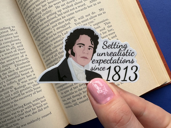 A cartoon of Mr Darcy from Pride and Prejudice with the text 'Setting unrealistic expectations since 1813'. The sticker is held above an open book on a blue background.