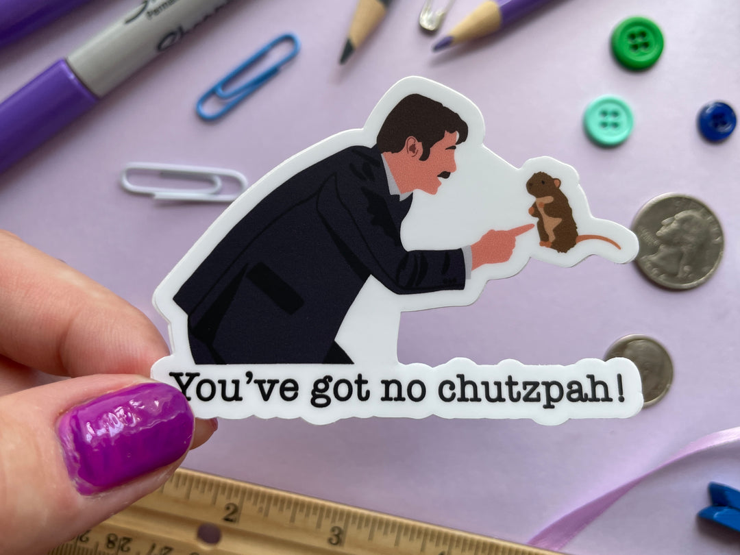 A sticker of a cartoon of comedian Mike Wozniak from the side pointing to a brown stuffed mole from the TV show Taskmaster with the words "you've got no chutzpah" beneath is held over a purple background covered in objects.