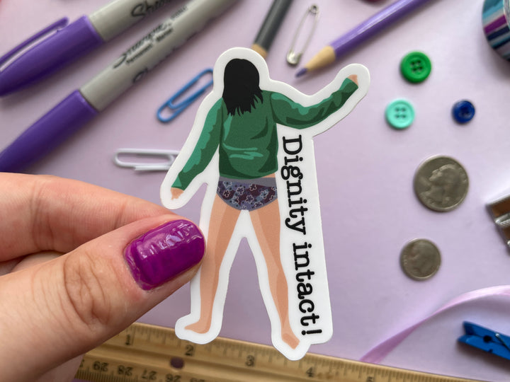 A sticker of a cartoon comedian Aisling Bea dancing away not wearing trousers from the TV show Taskmaster next to the text "Dignity Intact" is held over a purple background covered in objects.