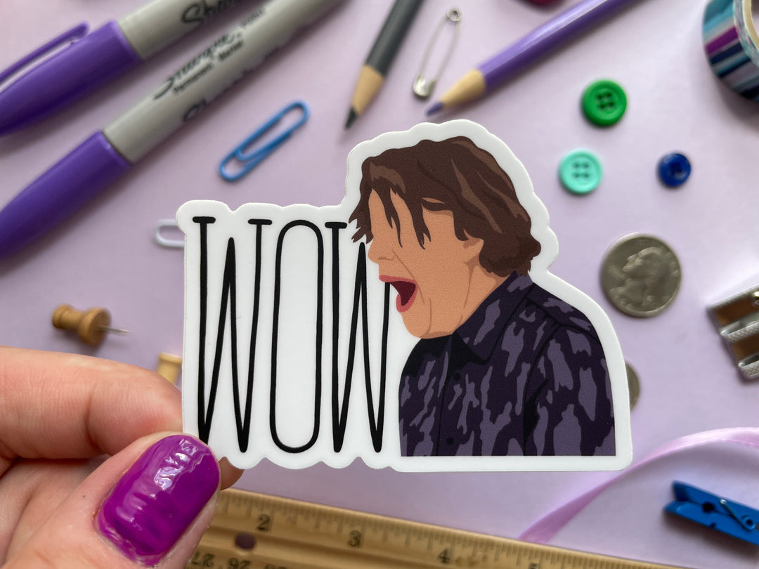 A sticker of a cartoon of comedia Alan Davies with his mouth opening shouting "Wow" from the TV show Taskmaster is held over a purple background covered in objects.