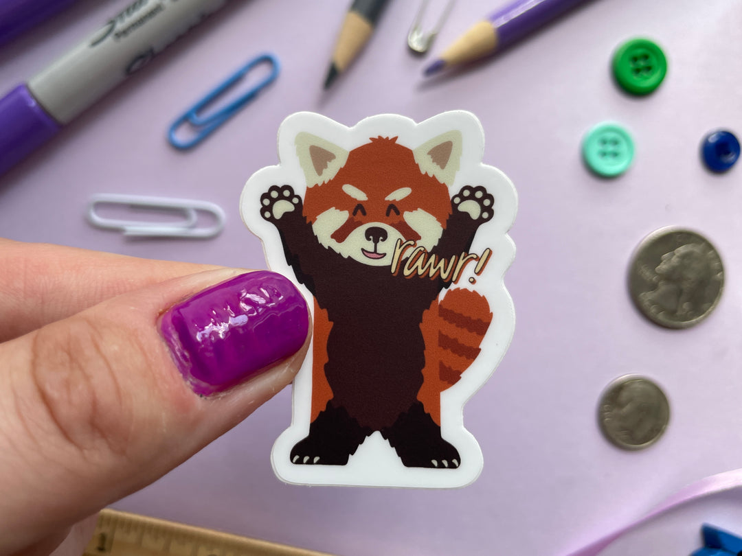 A sticker of a cartoon red panda standing on its hind legs with its arms raised and the text "rawr" next to its mouthis held over a purple background covered in objects.