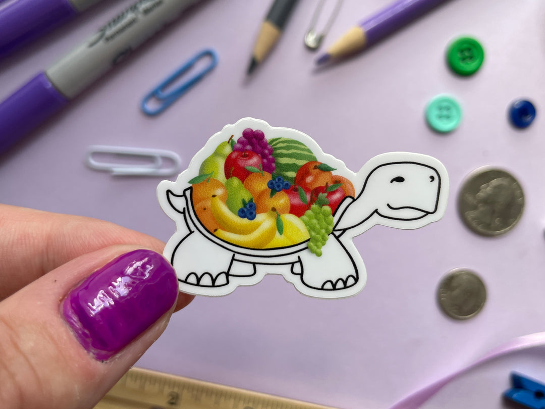 A sticker of a cartoon turtle drawn in black and white with a colorful shell of various fruits is held over a purple background covered in objects.