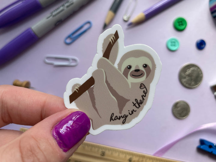 A sticker of a light brown sloth in profile hanging from a brand with the text "hang in there" and a heart over top is held over a purple background covered in objects.