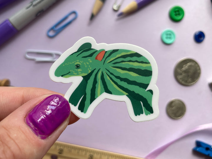 A sticker of a green cartoon tapir with watermelon stripes along its body is held over a purple background covered in objects.