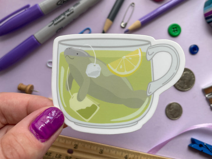 A sticker of a cup of green tea with a tea bag and slice of lemon with a grey manatee swimming in it is held over a purple background covered in objects.