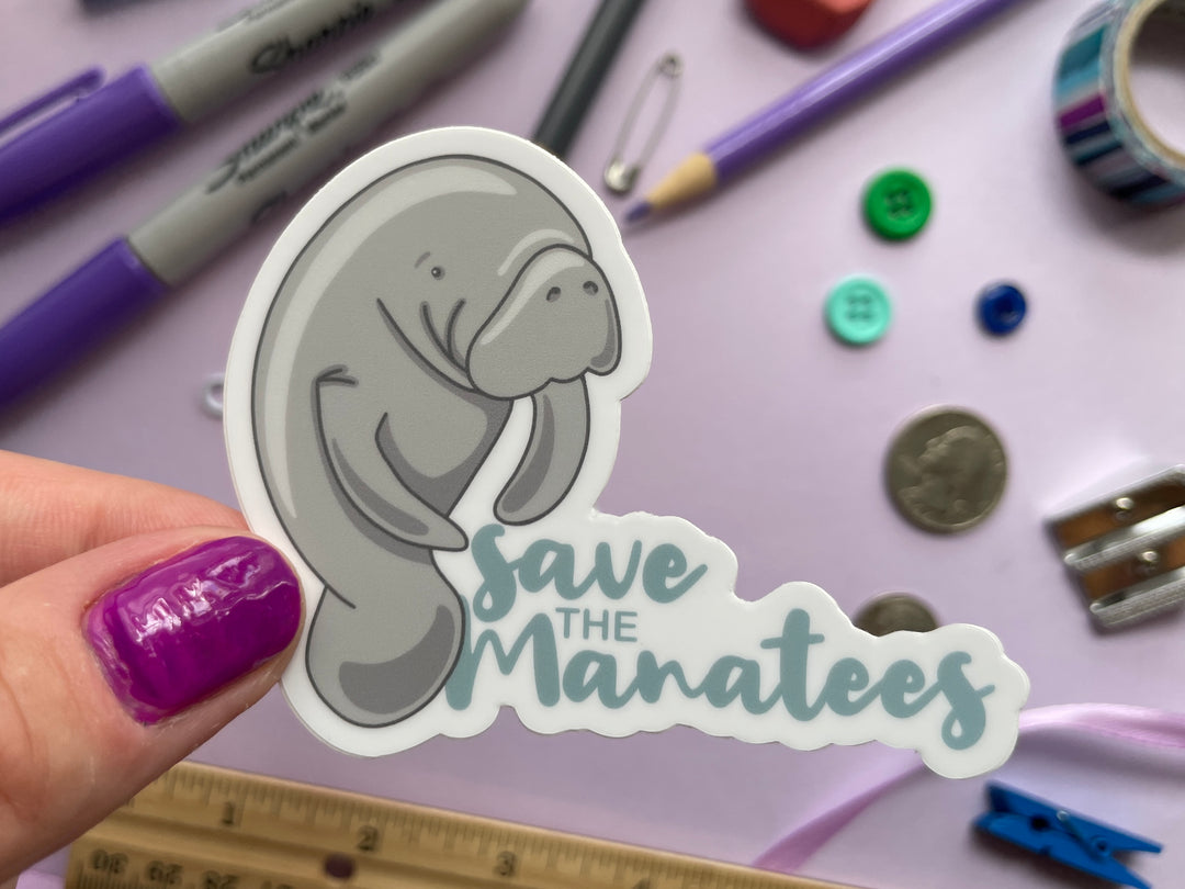 A sticker of a grey cartoon manatee standing of the size next to the light blue text "save the manatees" is held over a purple background covered in objects.