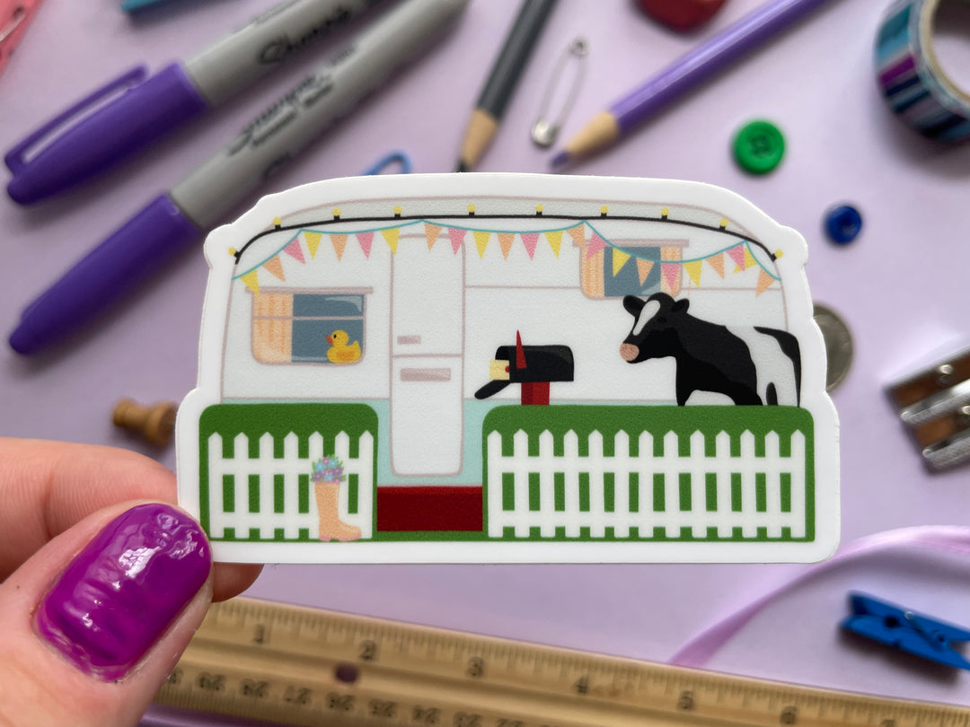 A sticker of the caravan from the TV show Taskmaster with a cow in the front yard and duck in the window is held over a purple background covered in objects.