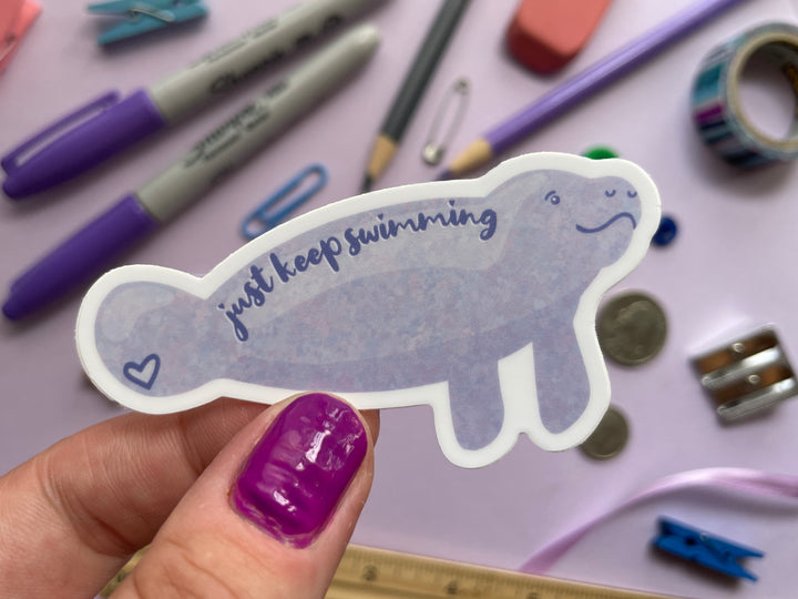 A sticker of a cartoon light purple smiling manatee shown from the side with the text "just keep swimming" over top is held over a purple background covered in objects.