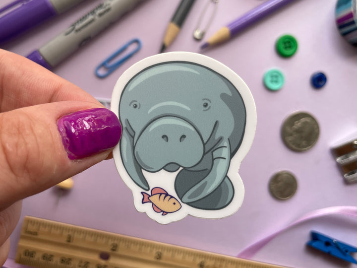 A sticker of a cartoon blue manatee head on with a yellow and orange fish beneath it is held over a purple background covered in objects.