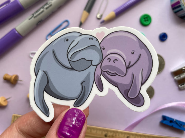 A sticker of a cartoon pair of manatees, one blue and one purple, with their heads together and a heart about them is held over a purple background covered in objects.