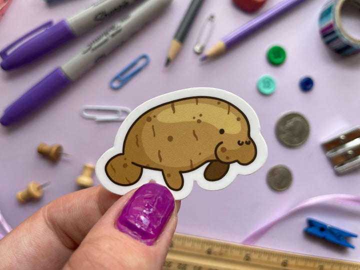 A sticker of a manatee shown in profile colored like a potato is held over a purple background covered in objects.