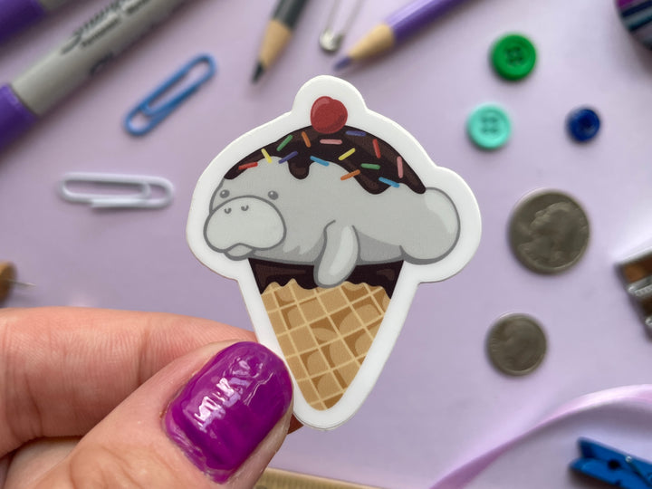 A sticker of a cartoon ice cream cone dipped in chocolate topped with a grey manatee with chocolate sauce, sprinkles, and a cherry is held over a purple background covered in objects.