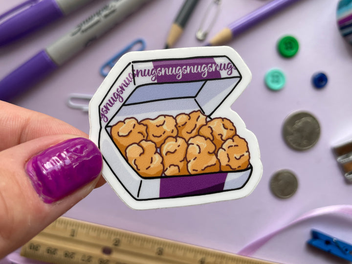 A sticker of a purple and white box of ten chicken nuggets with the word "nugs" repeated along the box is held over a purple background covered in objects.