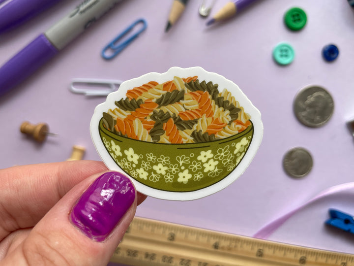 A sticker of a green bowl of orange, green, and cream colored spiraled rotini noodles is held over a purple background covered in objects.