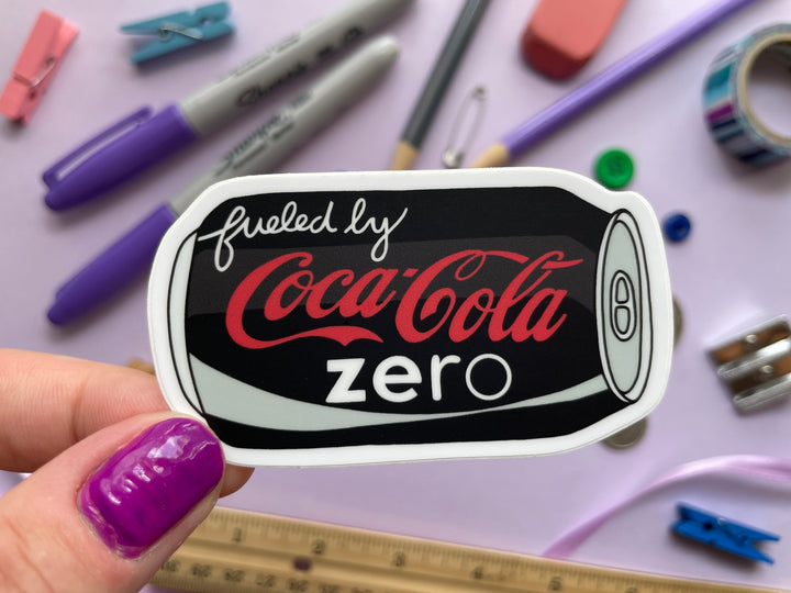 A sticker of a cartoon of a black can of Coke Zero on its side with the text "fueled by" in the top corner is held over a purple background covered in objects.