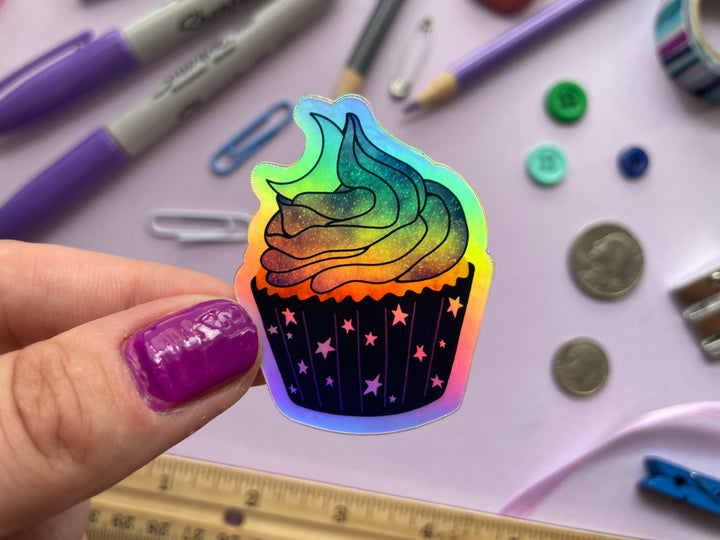 A holographic sticker of a cartoon of a cupcake with starry paper, purple and blue stardust frosting, and top with a crescent moon is held over a purple background covered in objects.