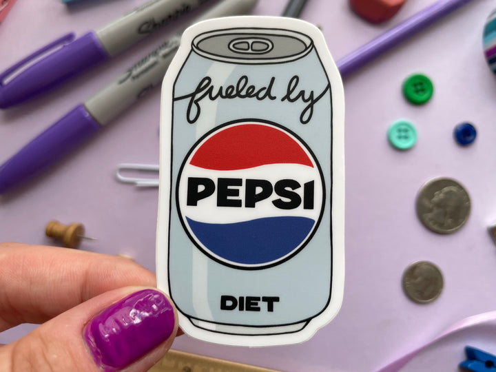 A sticker of a cartoon of a light blue can of Diet Pepsi with the text "fueled by" in the top corner is held over a purple background covered in objects.