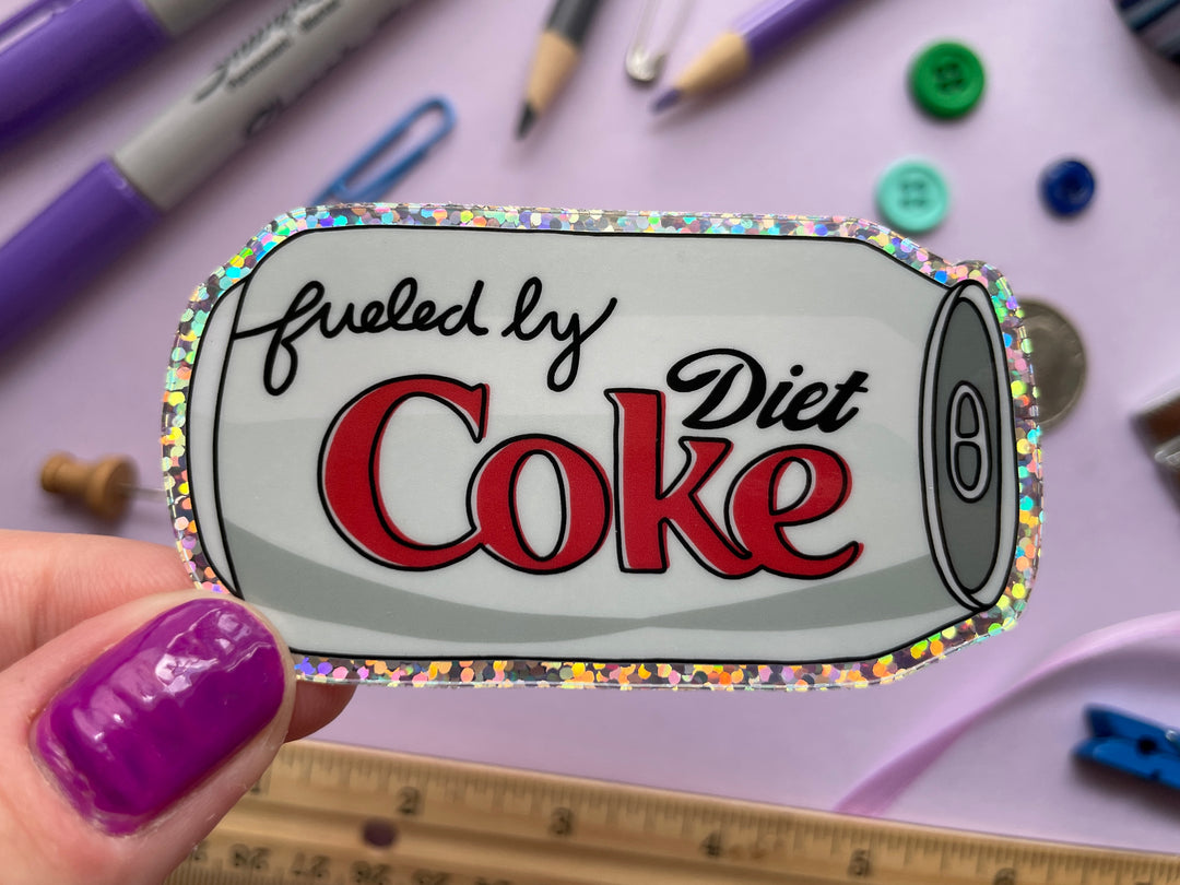 A glittery sticker of a cartoon of a silver can of Diet Coke on its side with the text "fueled by" in the top corner is held over a purple background covered in objects.