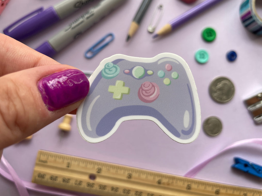 A sticker of a cartoon purple game controller with multi color buttons is held over a purple background covered in objects.