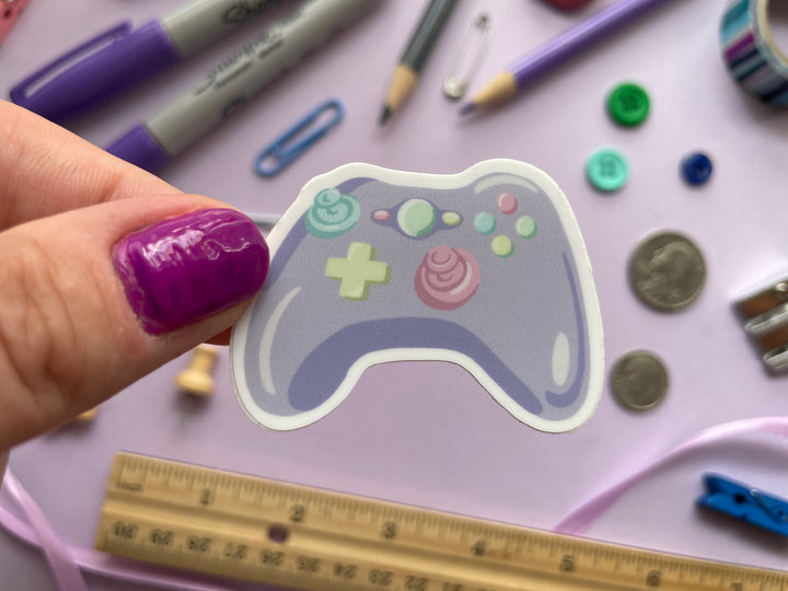 A sticker of a cartoon purple game controller with multi color buttons is held over a purple background covered in objects.