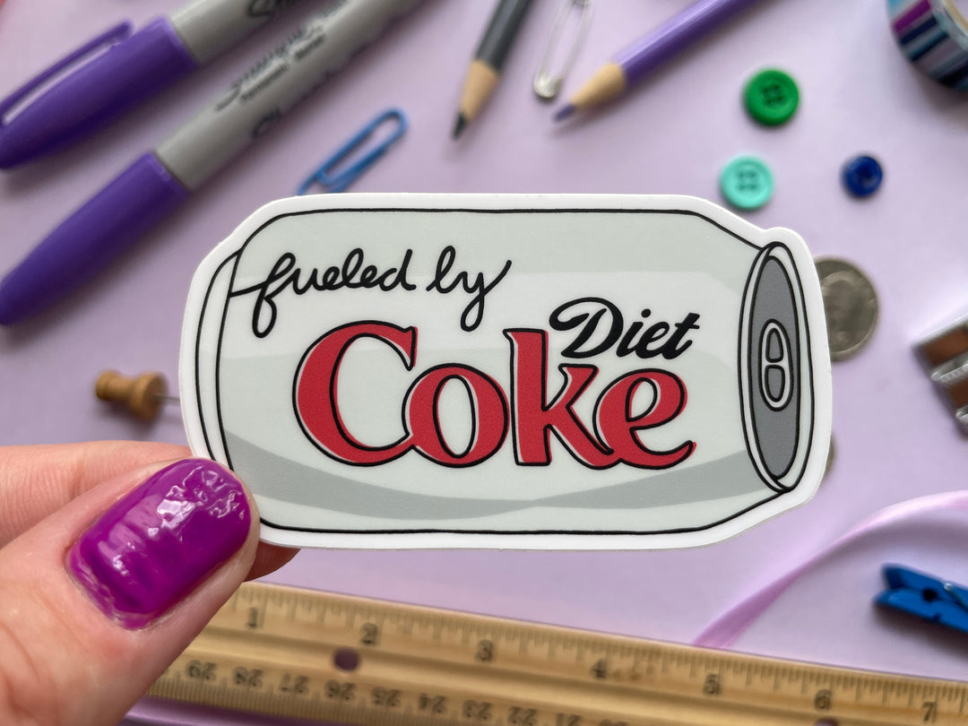 A sticker of a cartoon of a silver can of Diet Coke on its side with the text "fueled by" in the top corner is held over a purple background covered in objects.