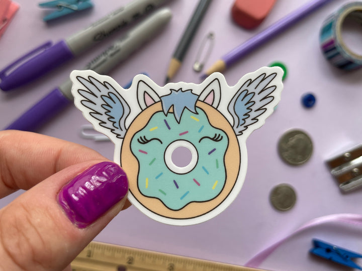 A sticker of a cartoon donut with blue frosting with blue feathered wings and horse ears and mane is held over a purple background covered in objects.