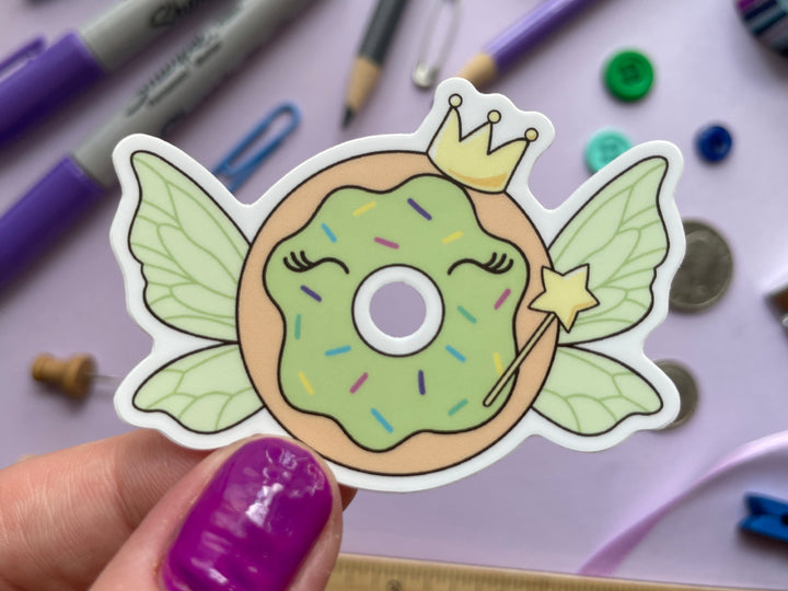 A sticker of a cartoon donut with green frosting with green fairy wings and a starry magic wand and crown is held over a purple background covered in objects.