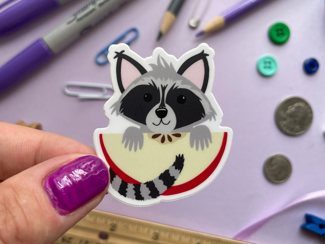 A sticker of a cartoon raccoon holding a large slice of red apple is held over a purple background covered in objects.