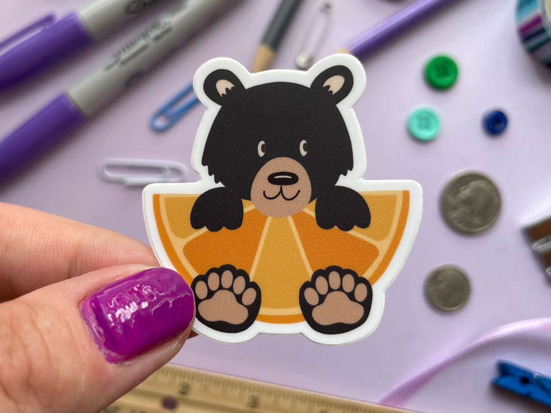 A sticker of a cartoon black bear holding a large orange fruit slice is held over a purple background covered in objects.