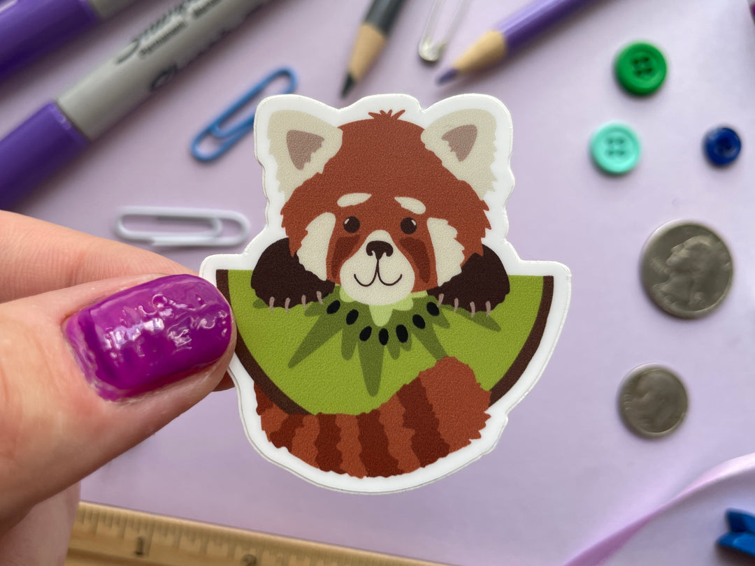 A sticker of a cartoon red panda holding a large slice of kiwi fruit is held over a purple background covered in objects.