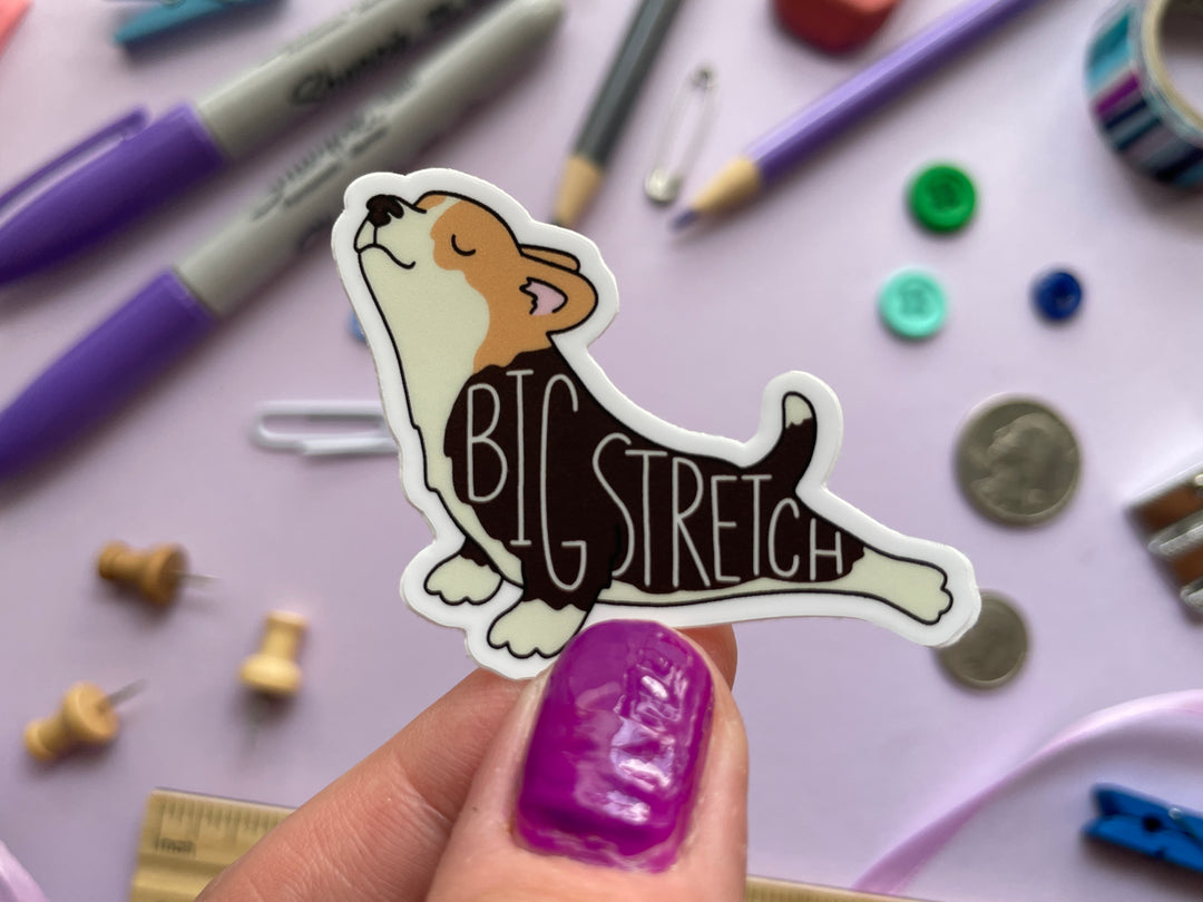 A sticker of a cartoon corgi stretching its back legs behind it with the text "big stretch" on top is held over a purple background covered in objects.