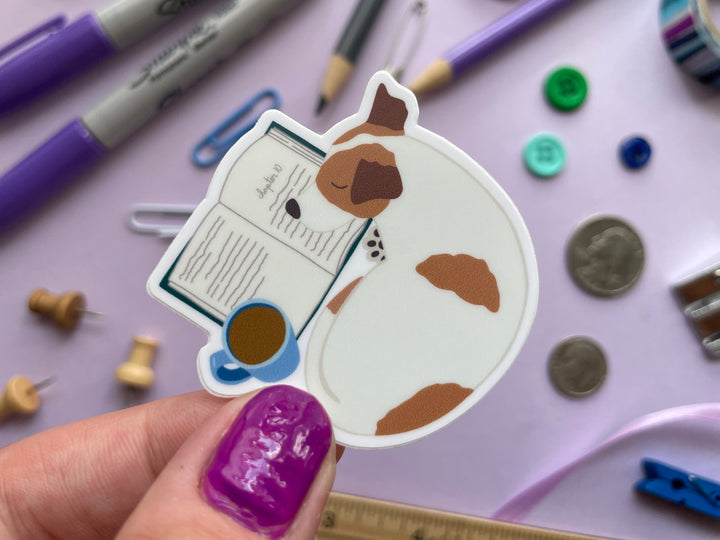 A sticker of a brown and white dog curled up next to an open box and mug of brown liquid is held over a purple background covered in objects.