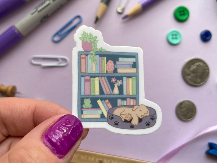 A sticker of a cartoon blue bookshelf with various items on the shelf and a plant on top with a sleeping puppy in a bed in front is held over a purple background covered in objects.