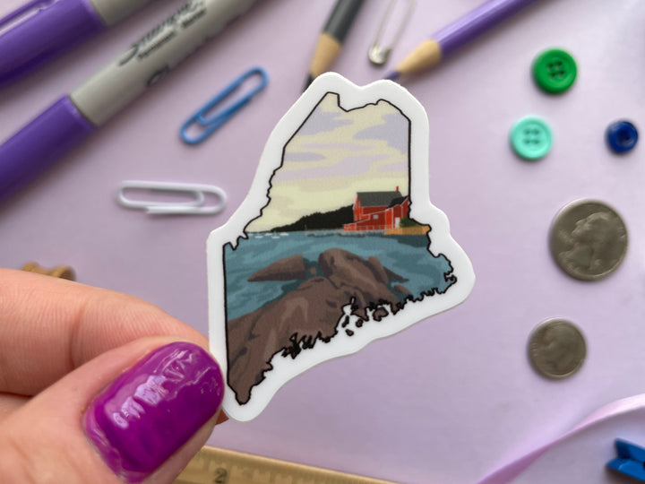 A sticker shaped liked the state of Maine depicting an ocean shoreline and red building is held over a purple background covered in objects.