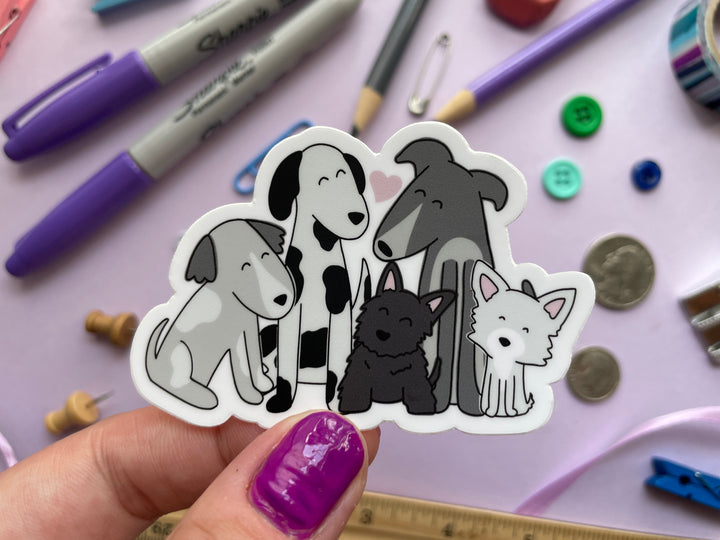 A sticker of a cartoon drawing of five dogs of different sizes in black and white sitting together with a pink heart above them is held over a purple background covered in objects.