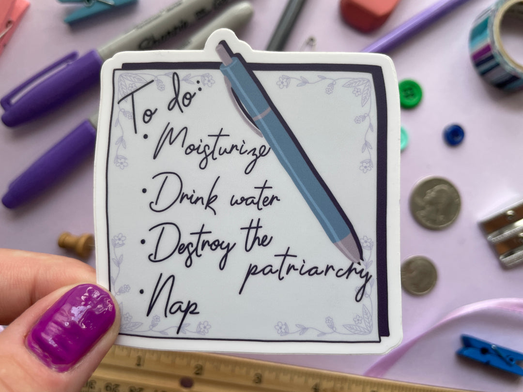 A sticker of a light purple square note with the text "To do: Moisturize, Drink water, Destroy the patriarchy, Nap" and a pen is held over a purple background covered in objects.