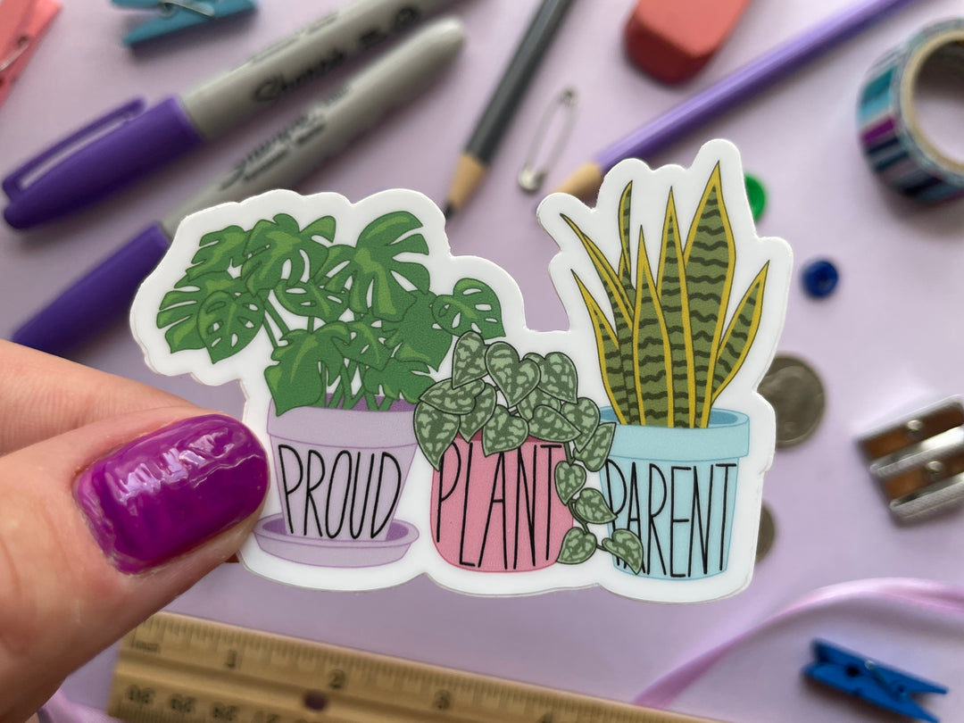 A sticker of a cartoon of three house plants in purple, pink, and blue planters with the text "proud plant parent" is held over a purple background covered in objects.