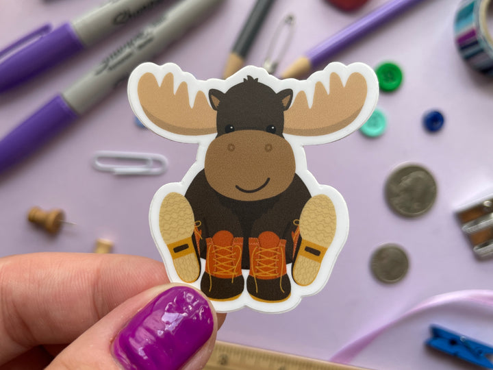 A sticker of a smiling cartoon moose sitting with orange and brown boots on its hooves is held over a purple background covered in objects.